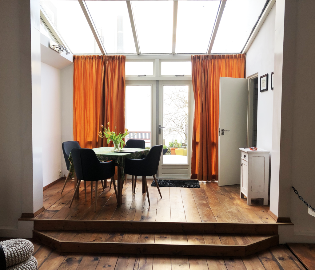 A photo of a table and chairs inside an apartment with a broad skylight and orange curtains framing sliding doors that lead to a patio