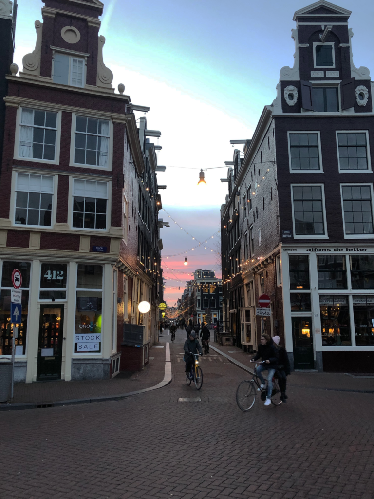 A photo of leaning Amsterdam canal houses on either side of a narrow cobblestone street with cyclists, in front of a pink-and-blue sunset-streaked sky