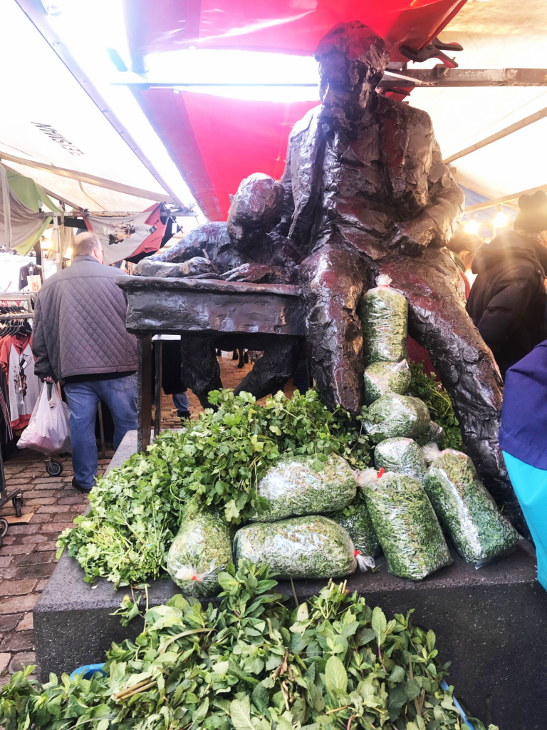 A photo of a modern statue of a man helping a boy with work at a desk, surrounded by bags of parsley in a bustling farmers market