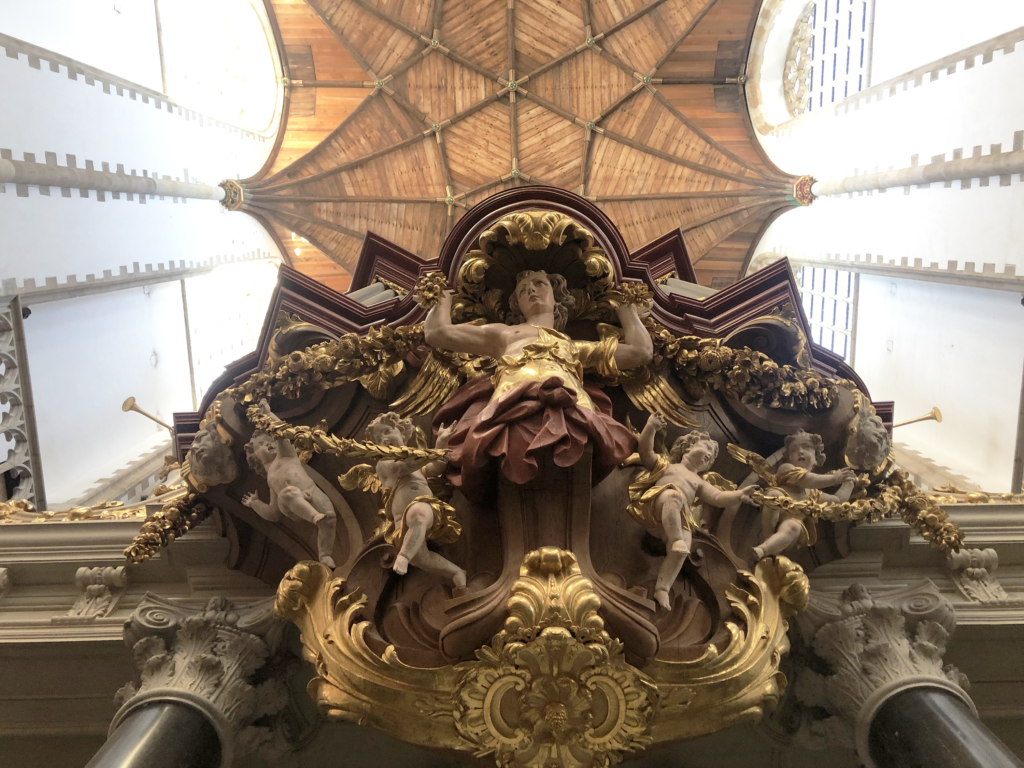 A photo looking up at painted wood carvings of cherubs and gilded garlands, below a ceiling made of light-brown wooden rafters