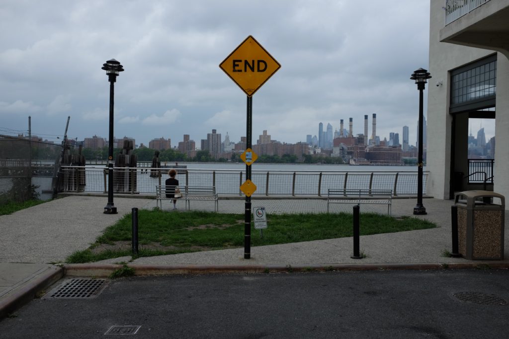 a yellow sign reading "END" in front of a waterfront, with the New York skyline beyond it