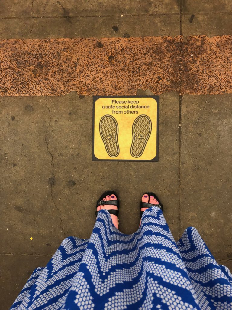 feet in sandals below a blue-and-white-dress next to a social distancing floor sticker