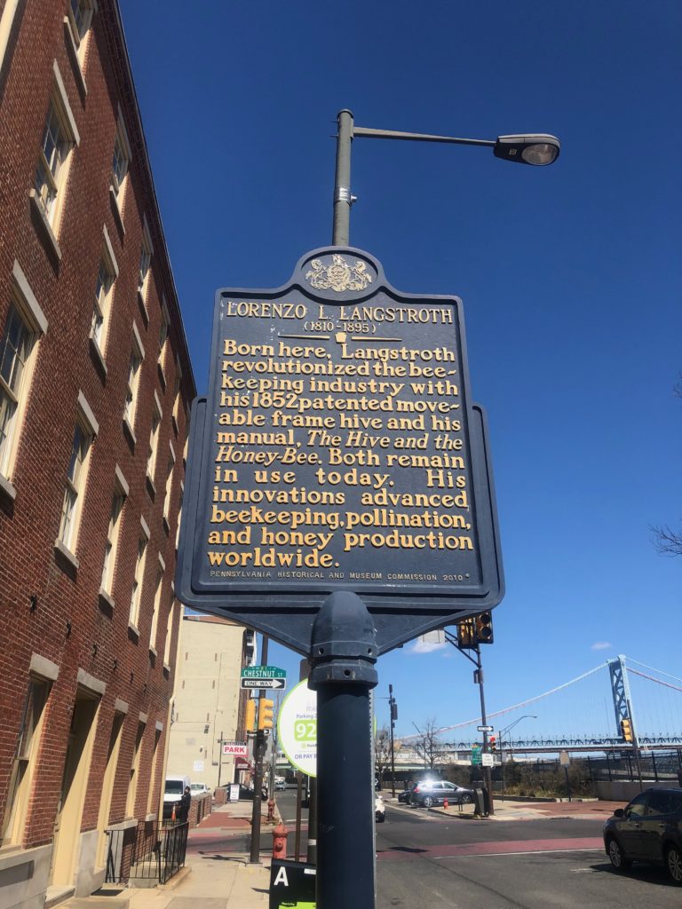 A blue historical sign for Lorzeno L Langstroth, 1810-1895, reading, "Born here, Langstroth revolutionized the beekeeping industy with his 1852 patented movable frame hive and his manual The Hive and the Honey-Bee. Both remain in use today. His innovations advanced beekeeping, pollination, and honey production worldwide.