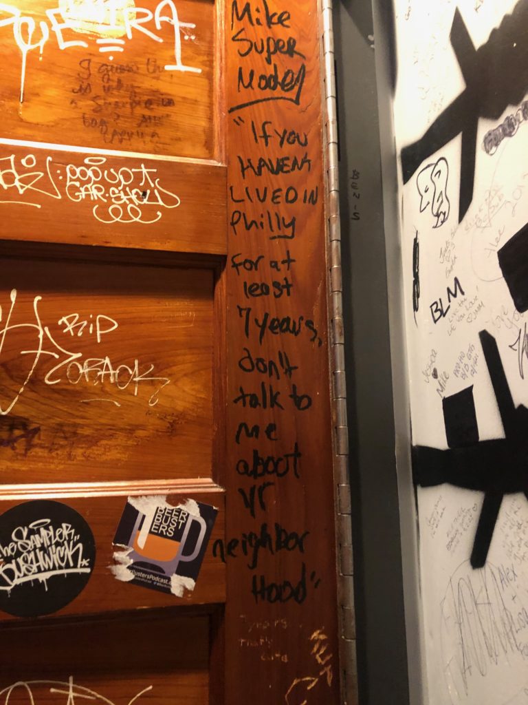 A wooden door covered in stickers and scribbles, with "If you haven't lived in Philly for at least 7 years, don't talk to me about yr neighborhood" written in black on it