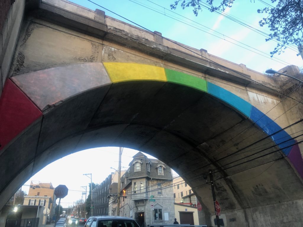 A cement bridge with a rainbow drawn along its inner arch. Beneath it is a few of cars and houses.