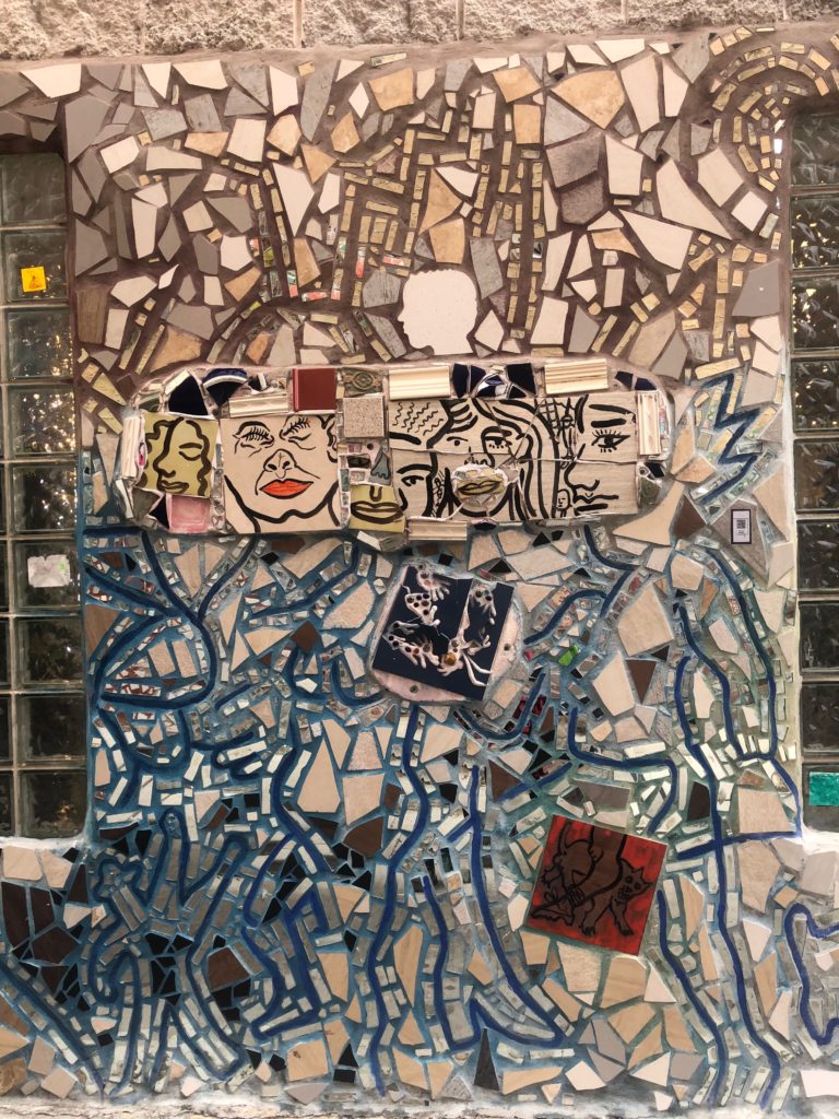 A tile wall with faces drawn on some of the tile pieces. Others are in shades of shite, cream, pink, and blue, and some are mirrored.