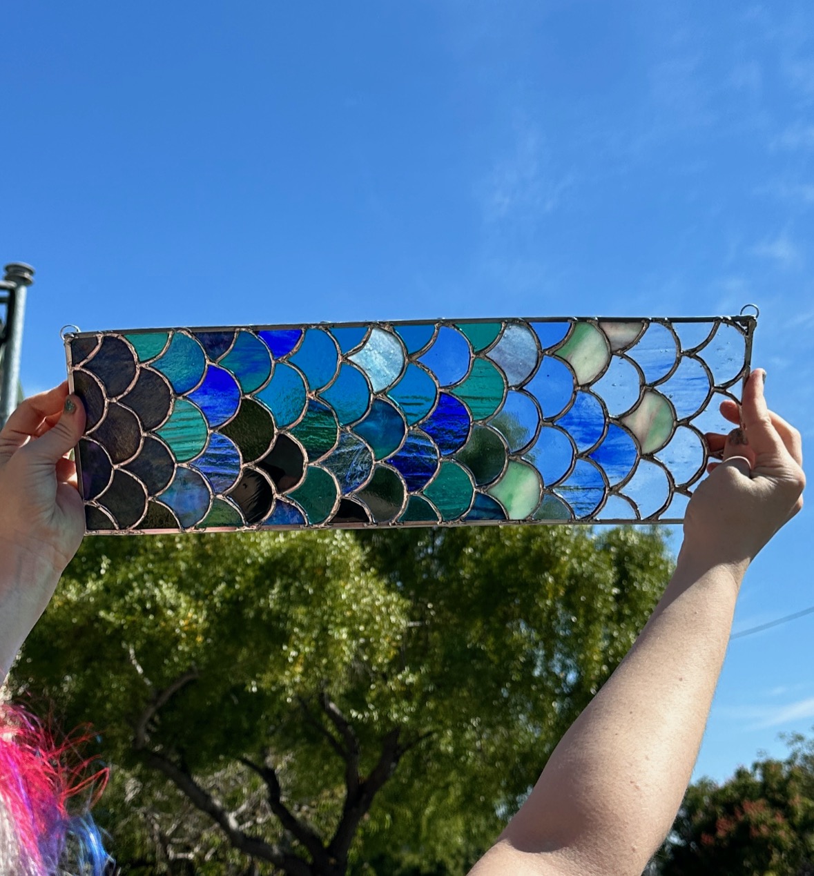 A white woman's arms hold a rectangular stained glass pane up against the sky and trees. The glass is many shades of blue, green, and clear, done in the shape of fish scales.