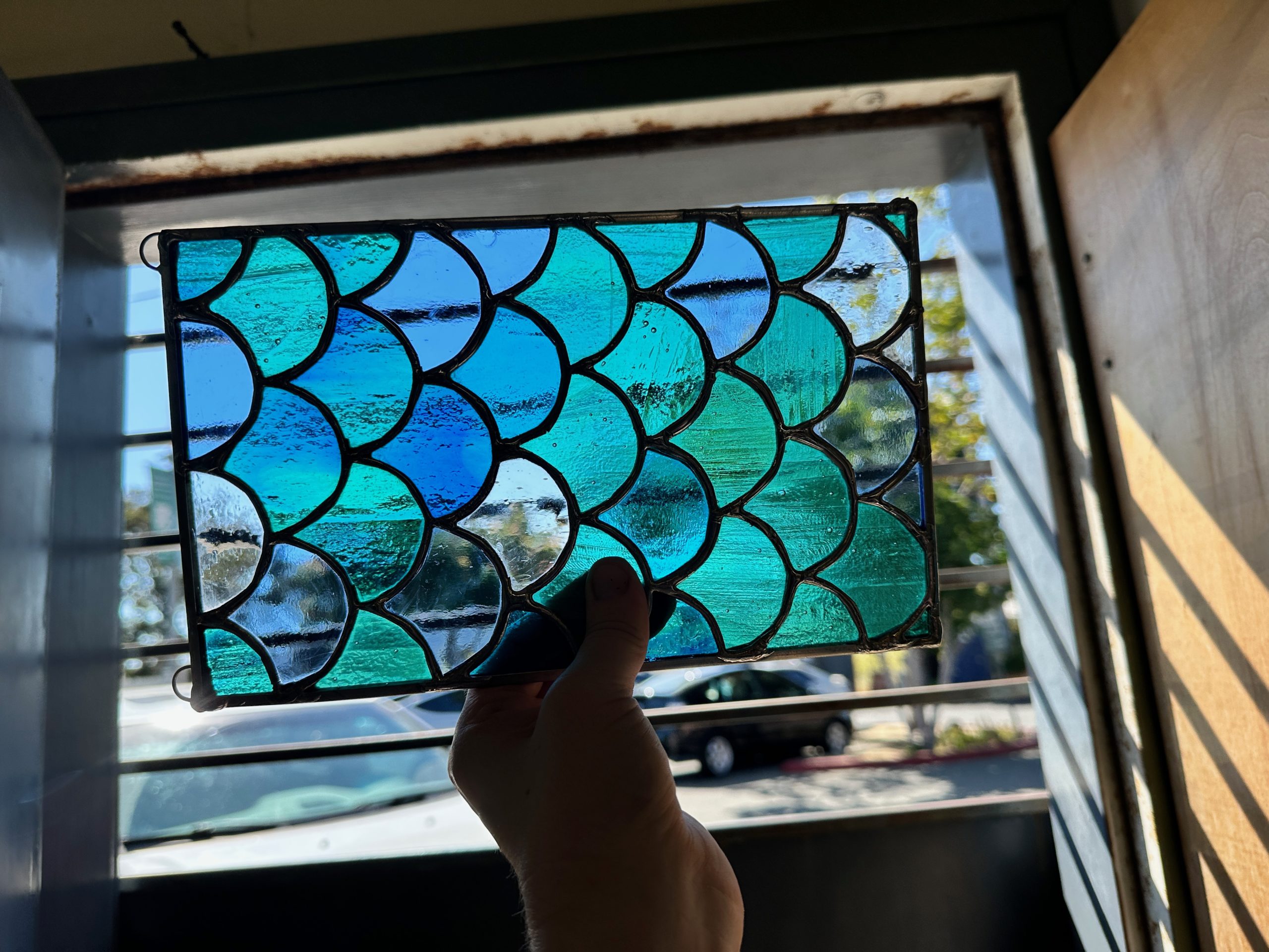 A stained glass piece with a fish-scale pattern in blues and greens, held up to the light by a white person's hand