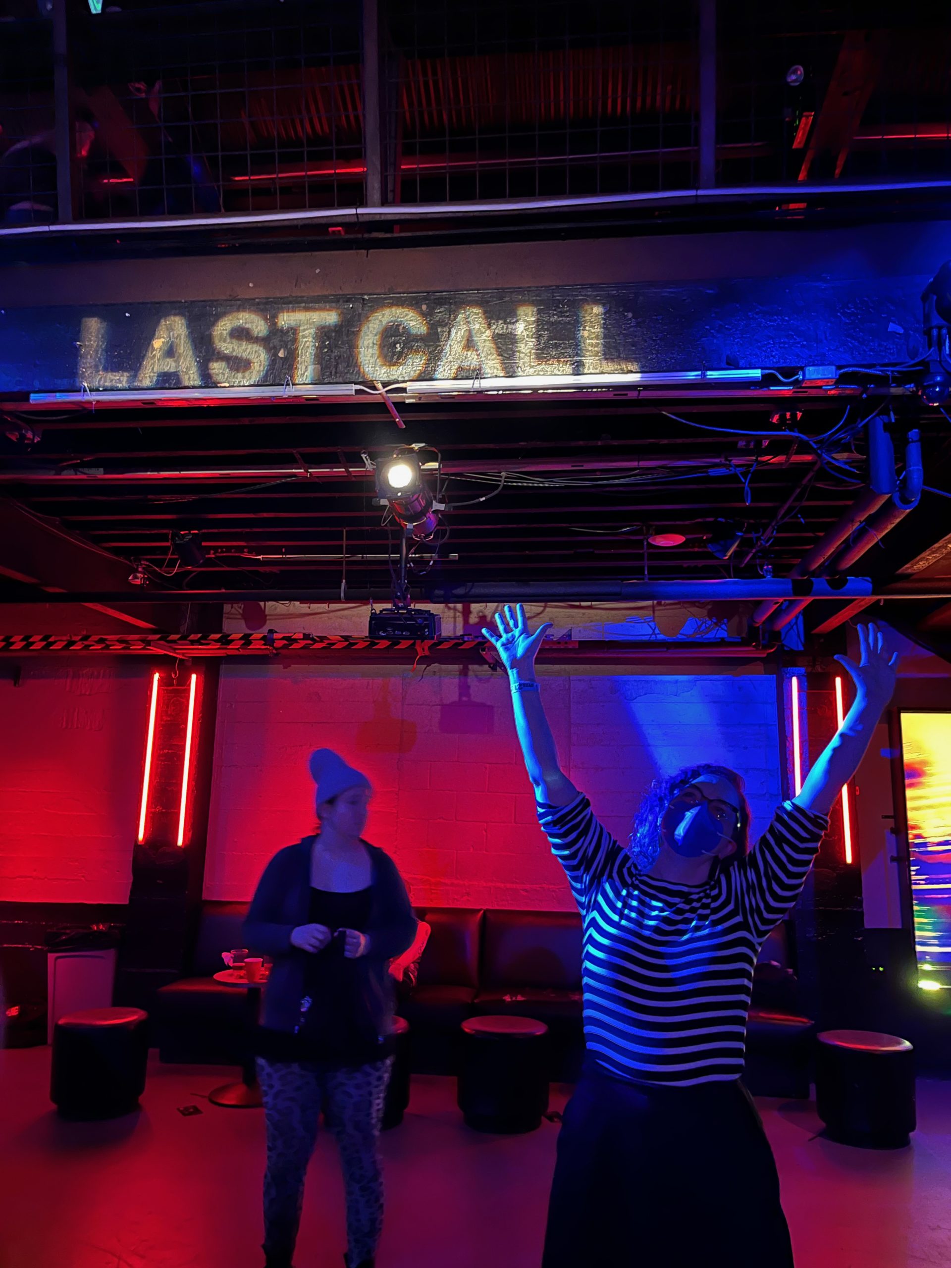 The author, hands in the air, in a black-and-white striped shirt and black skirt, in a red-lit bar with LAST CALL in light against a wall overhead, an unwitting bystander behind her who probably wishes to be excluded from this narrative