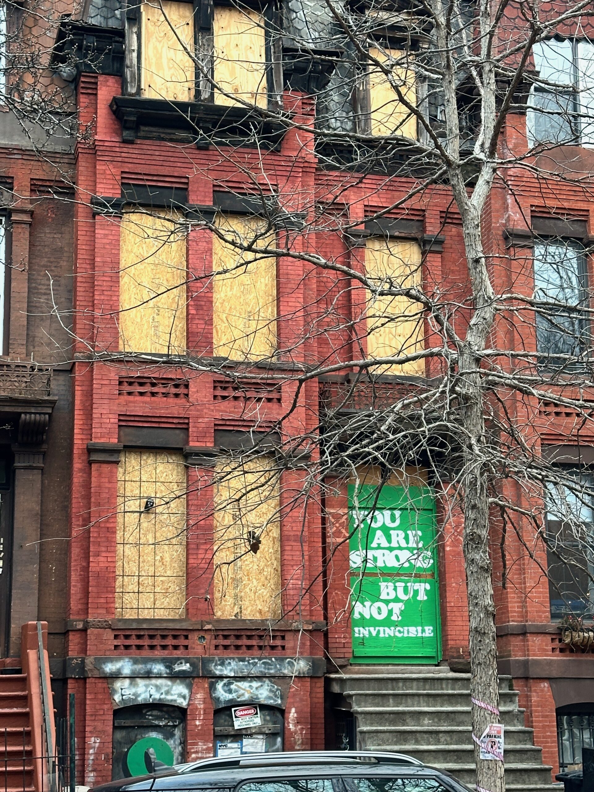 A brick townhouse in Brooklyn with windows covered in raw plywood. The front door is also covered in plywood and boards, painted kelly green, with white lettering that says, "You are strong but not invincible." It's winter, and a tree with bare branches is in the foreground.