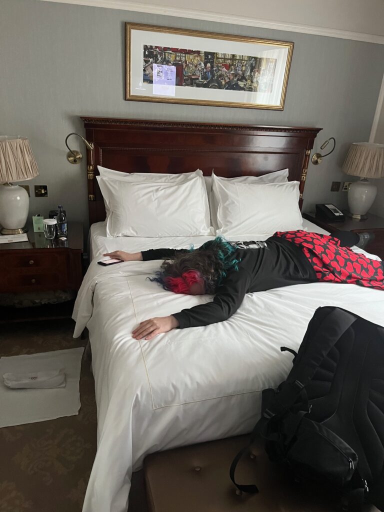 A white woman in a black hoodie and black skirt printed with red hearts lies face down, arms over her head on a hotel room bed with nice white linens