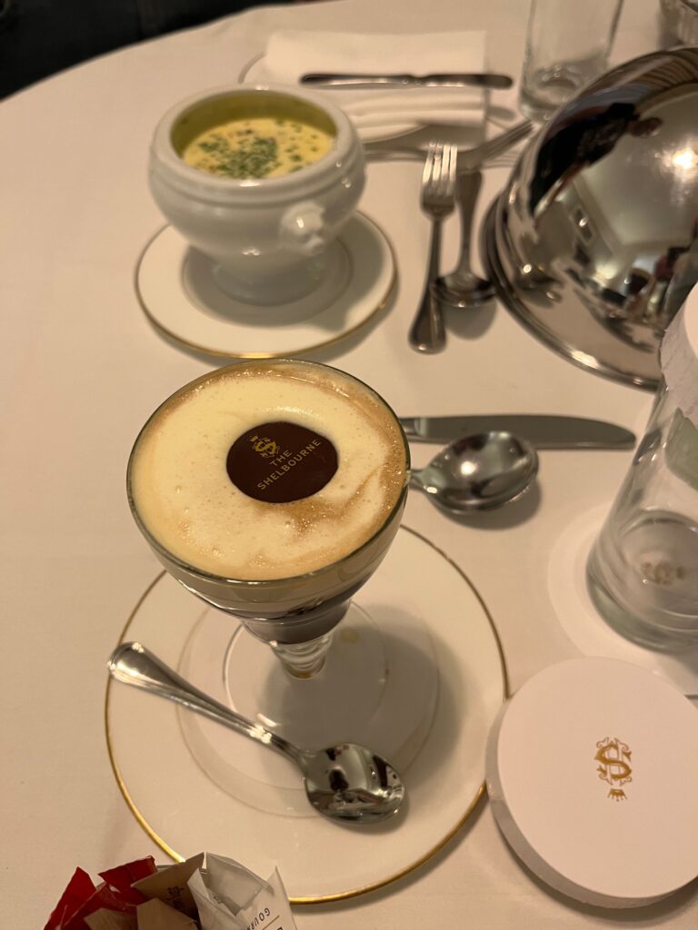 A table with a white tablecloth holes an Irish coffee with a chocolate disc on top, a silver spoon next to it. Other silverware and dishes are out of focus in the background.