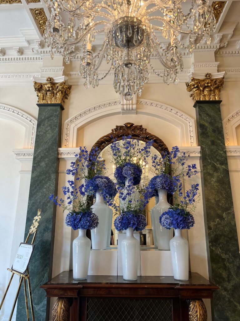 An elaborate space in a hotel lobby with marble columns and gilded molding surrounding a polished wood-and-marble table with six white vases filled with blue-purple hydrangea and other flowers